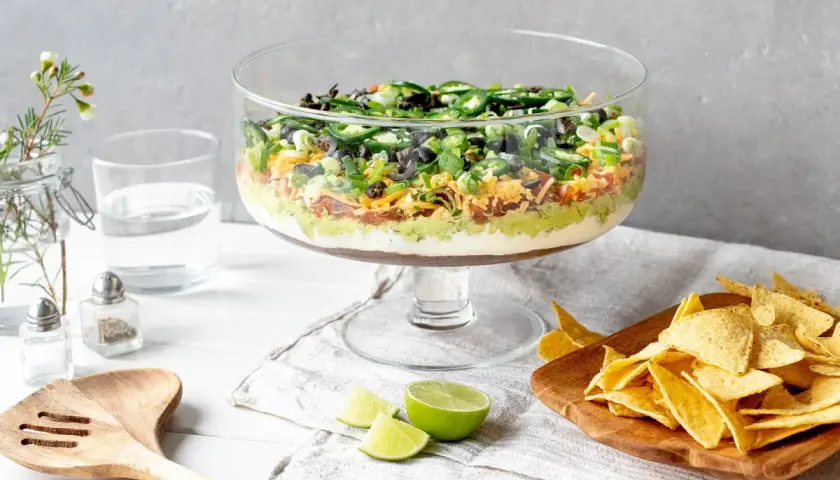 Schicht für Schicht ein Genuss: Ein bunter 7-Layer-Dip in einer Glasvitrine, bereit zum Dippen mit Tortilla-Chips.