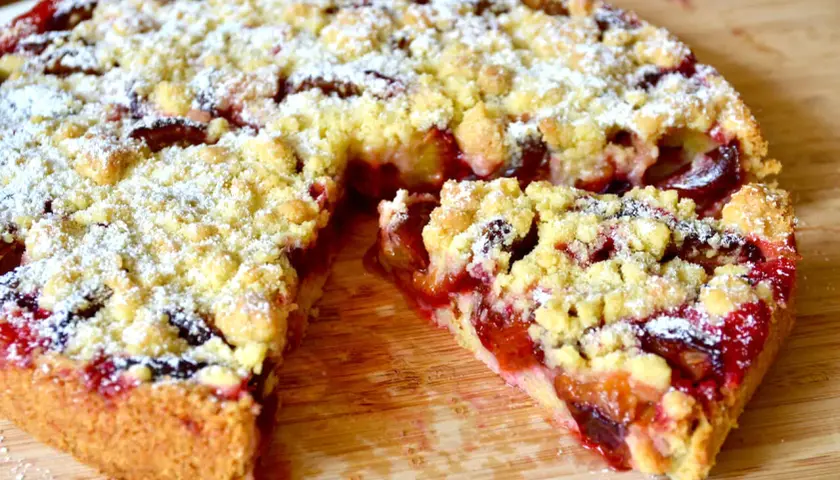 Omas Pflaumenkuchen aus Hefeteig in Springform, mit Streuseln und Puderzucker bestäubt. Ein Stück ist herausgeschnitten.