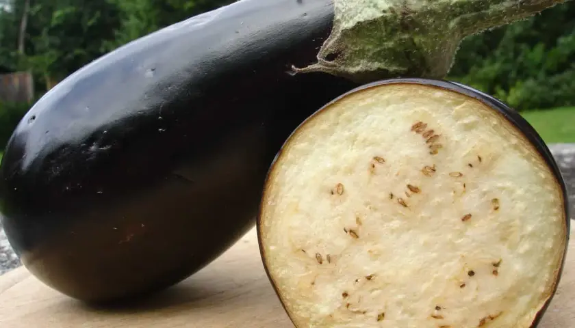 Eine glänzende, dunkle Aubergine liegt neben einem aufgeschnittenen Exemplar. Im Inneren der Aubergine sind viele kleine, braune Kerne zu sehen, die wie schwarze Punkte wirken.