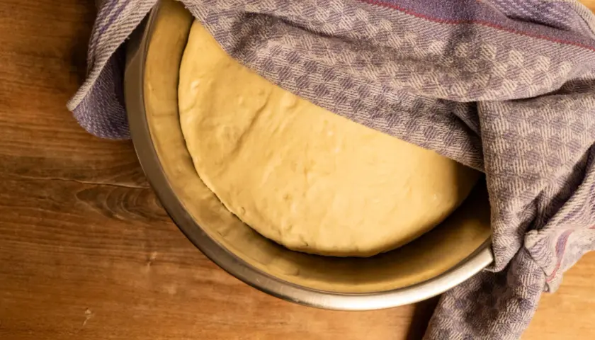 Frischer, aufgegangener Teig für pizzateig selber machen ruht in einer Schüssel unter einem karierten Tuch.