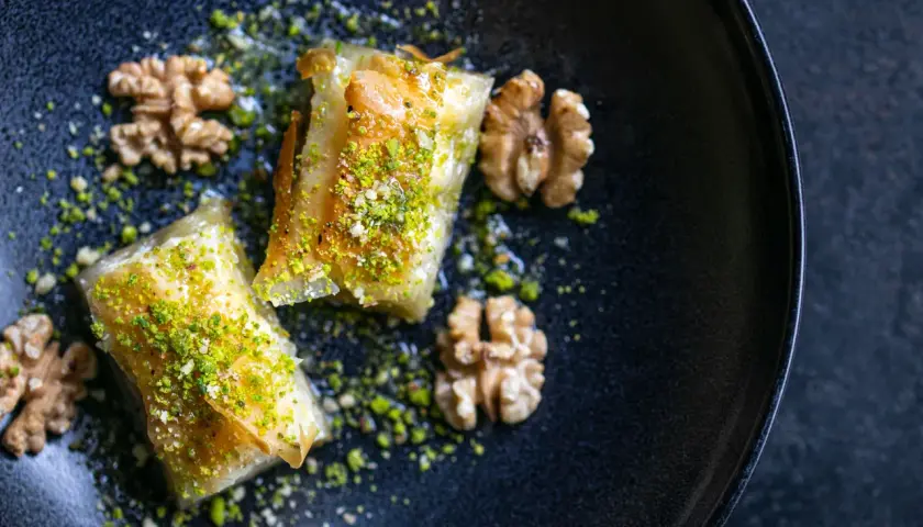 Zwei goldbraune Baklava-Stücke, bestreut mit gehackten Pistazien und Nüssen, auf einem dunklen Teller. Ein köstliches Baklava Rezept.