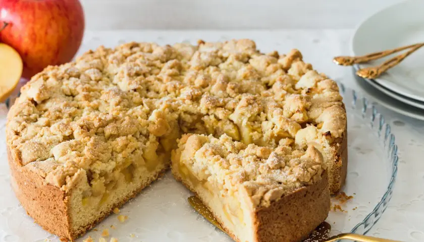 Ein saftiger, bester Apfelkuchen mit Streusel, bereit zum Servieren. Daneben liegen rote Äpfel.