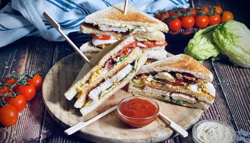 Drei saftige Club Sandwiches mit Hähnchen, Speck, Ei und Tomaten, serviert mit Ketchup und Mayo.