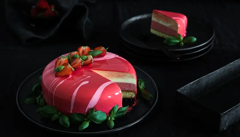 Eine Mousse Torte mit glänzendem Spiegelglasur, dekoriert mit Erdbeeren und Basilikum. Ein Stück ist auf einem Teller angerichtet.
