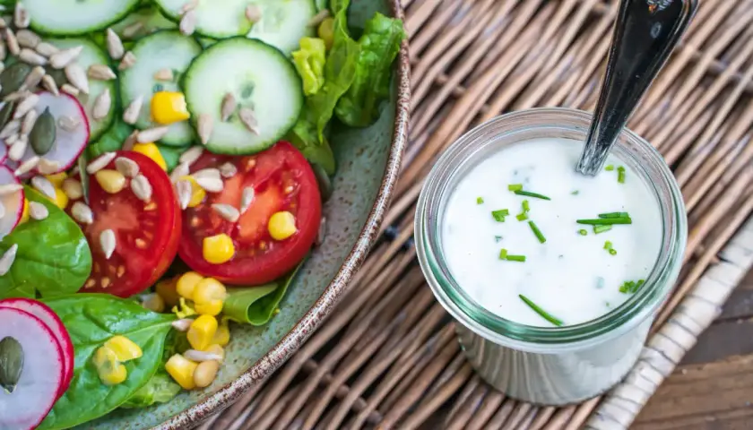 Frischer Salat mit Tomaten, Gurken und Mais neben einem Glas mit selbstgemachtem Joghurt-Dressing, garniert mit Schnittlauch.