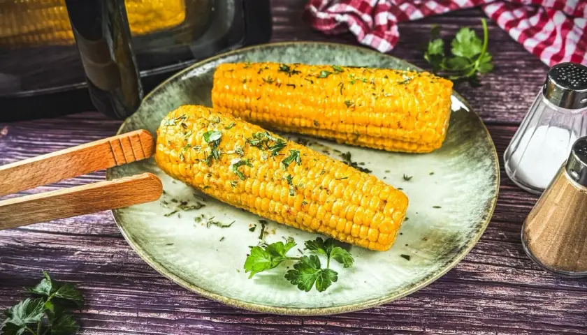 Zwei goldgelbe Maiskolben, frisch aus dem Airfryer, mit Kräutern bestreut, liegen auf einem Teller. Daneben Salz- und Pfefferstreuer.