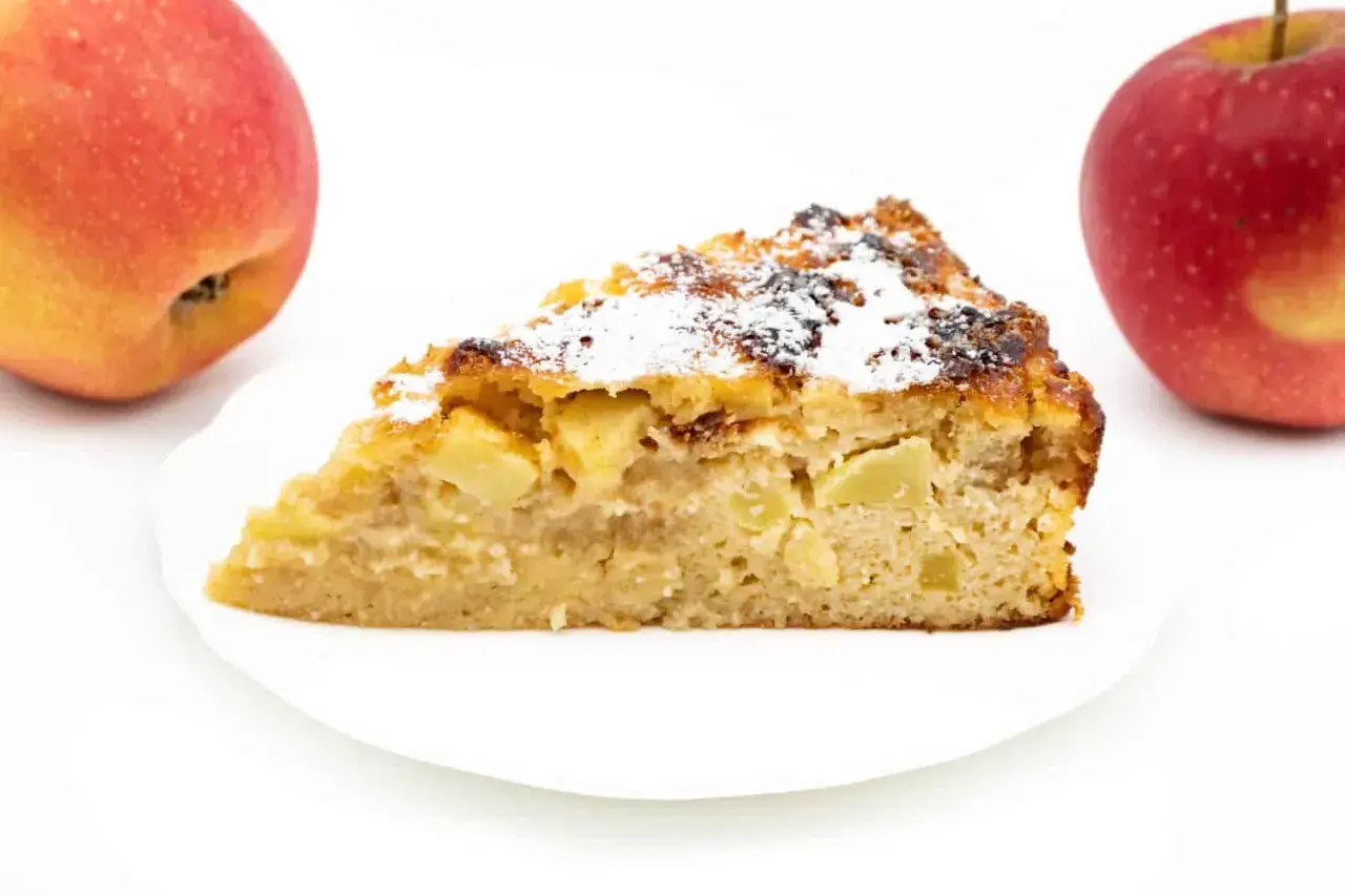 Ein Stück Apfelkuchen ohne Zucker mit Quark, bestäubt mit Puderzucker. Zwei Äpfel liegen daneben.
