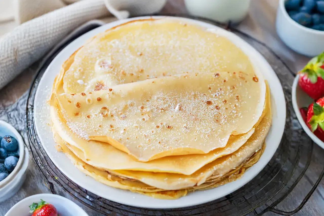 Ein Stapel goldbrauner Pfannkuchen, bestäubt mit Puderzucker. Daneben Schüsseln mit Blaubeeren und Erdbeeren. Ein sehr gutes Pfannkuchen Rezept für den Genuss.