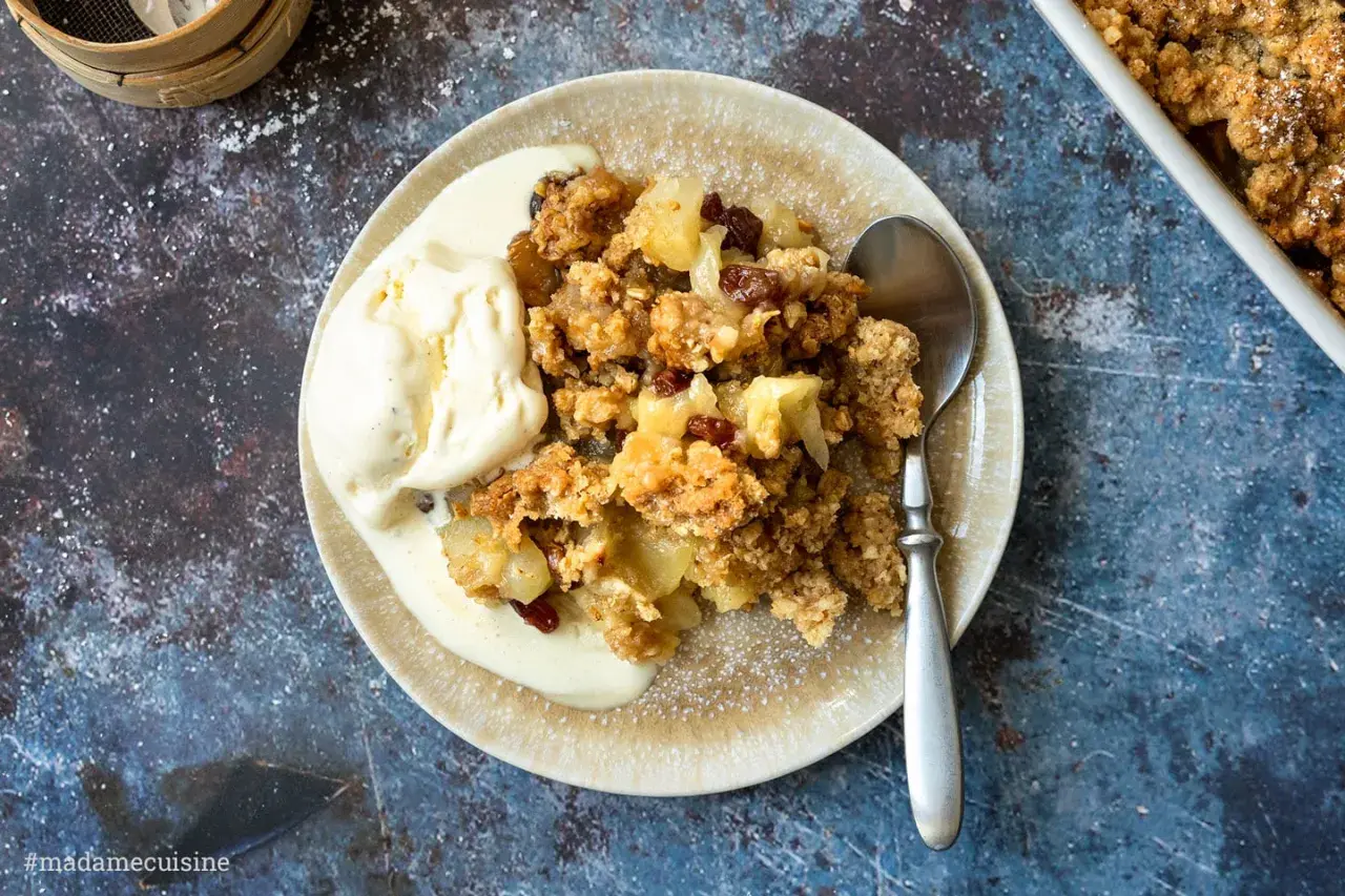 Ein Teller mit warmem Apfelcrumble und einer Kugel cremigem Vanilleeis, bestäubt mit Puderzucker.