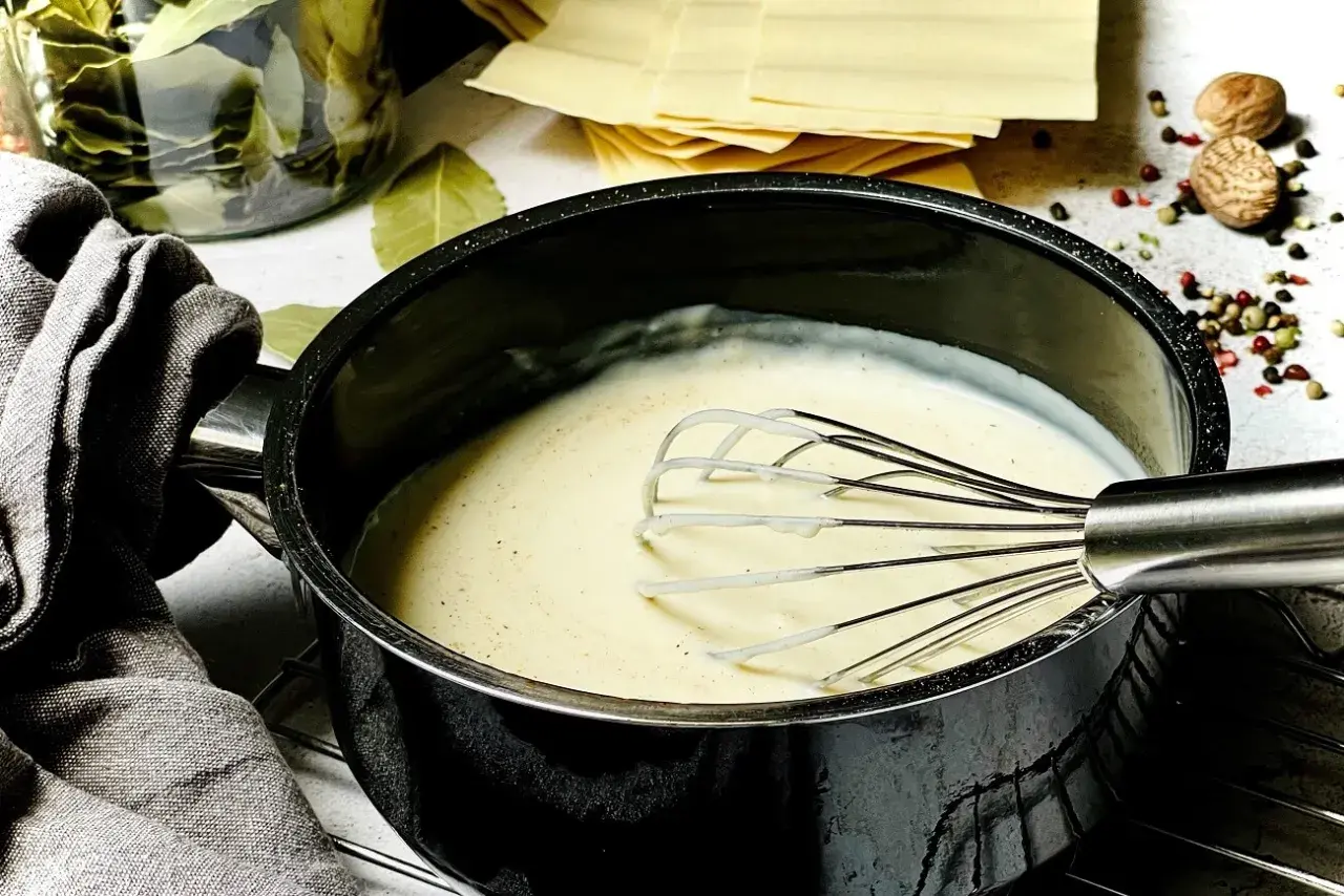 Cremige Béchamelsauce wird in einem Topf mit einem Schneebesen verrührt. Daneben liegen Lasagneplatten, Lorbeerblätter und Gewürze.