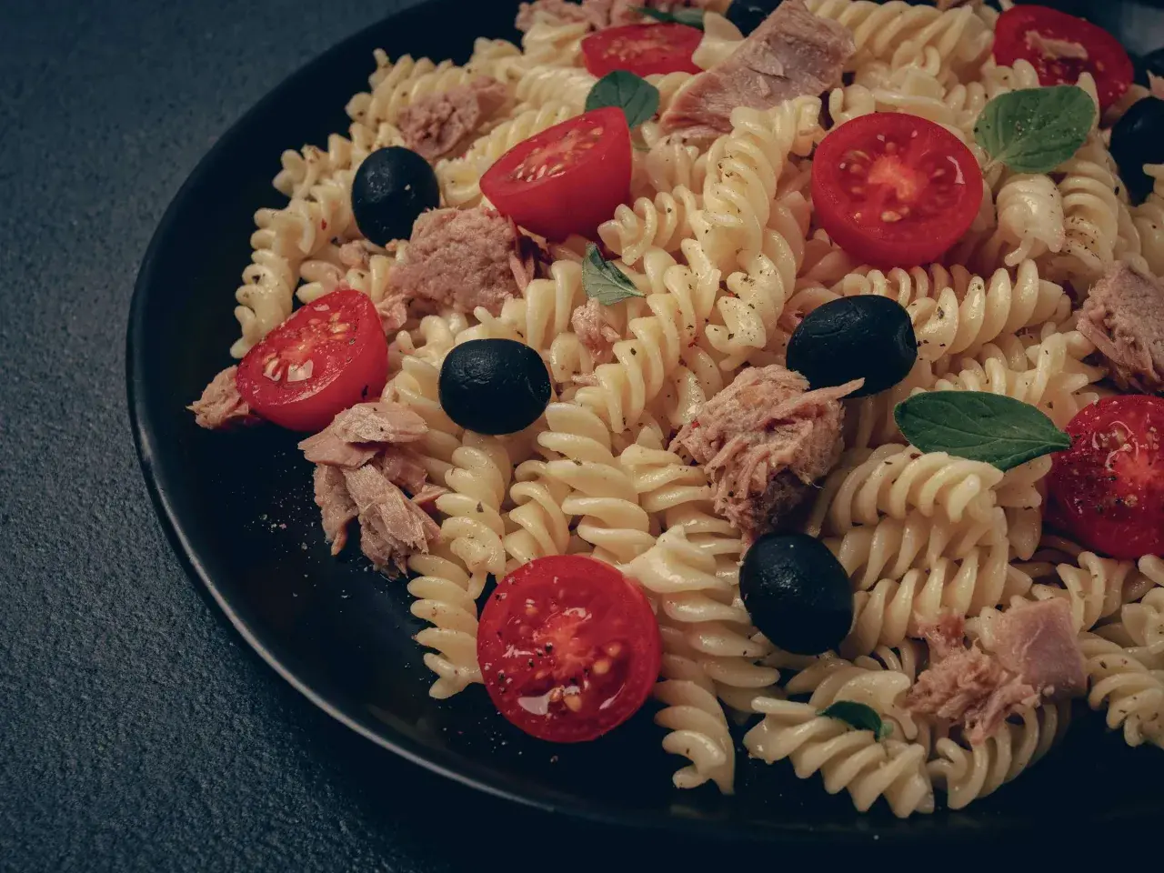 Ein köstlicher italienischer Nudelsalat mit Fusilli, Thunfisch, Kirschtomaten und schwarzen Oliven auf einem schwarzen Teller.