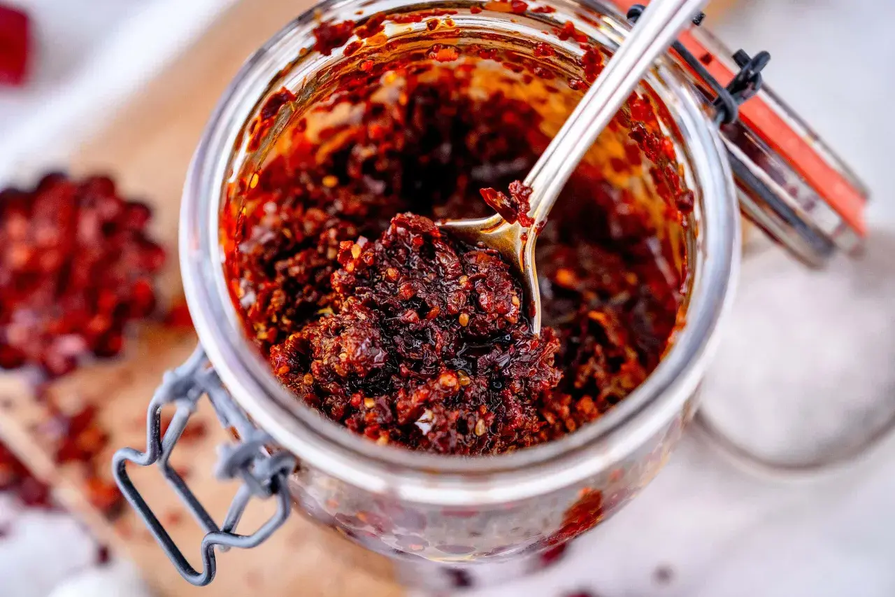 Ein Glas mit selbstgemachtem, würzigem Chiliöl. Ein Löffel hebt die glänzende, rote Masse hervor, perfekt für jedes crispy chili oil rezept.