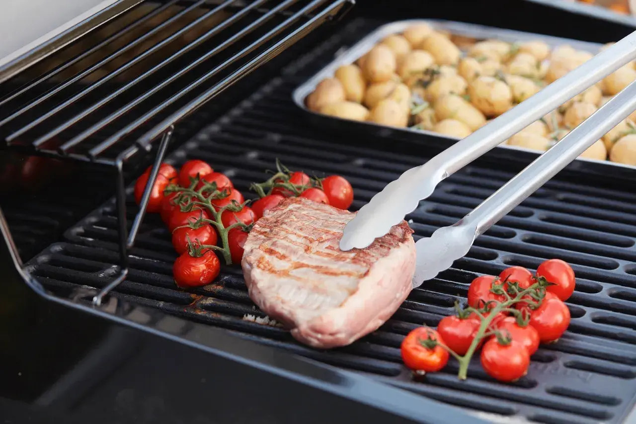 Duroc secreto zubereitung: Ein saftiges Steak wird mit Kirschtomaten auf dem Grill zubereitet. Daneben liegen Kartoffeln in einer Schale.