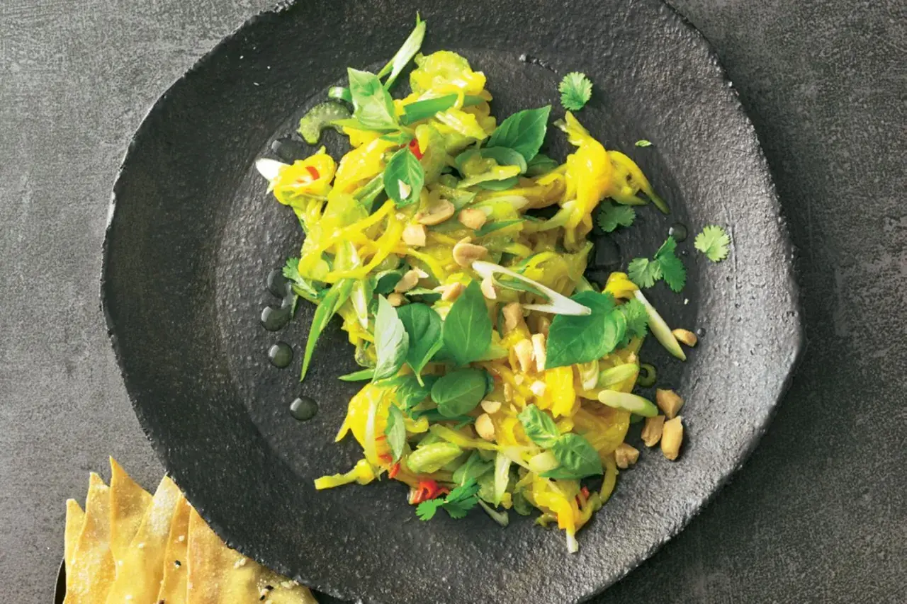 Ein bunter, frischer Mango Salat Thai mit Erdnüssen und Kräutern auf einem dunklen Teller.