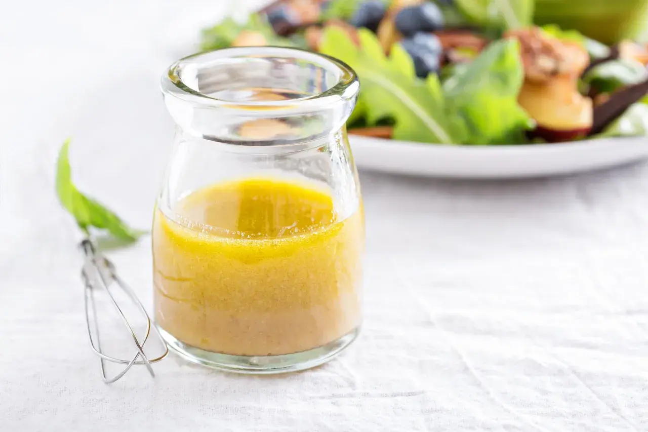 Ein Glas mit gelber Vinaigrette, daneben ein Schneebesen. Ein einfaches vinaigrette rezept für frische Salate.