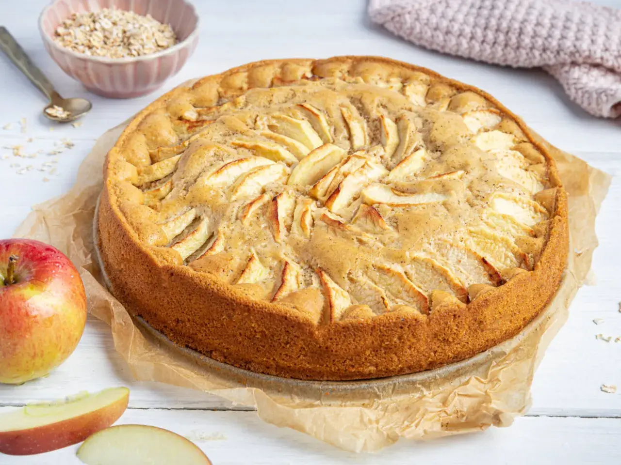 Ein goldbrauner, saftiger apfelkuchen ohne ei, dekoriert mit Apfelscheiben. Daneben liegen frische Äpfel und eine Schüssel Haferflocken.
