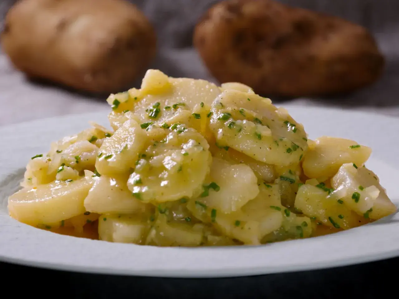 Ein Teller mit klassischem deutschen Kartoffelsalat, garniert mit frischen Kräutern. Im Hintergrund sind rohe Kartoffeln zu sehen.