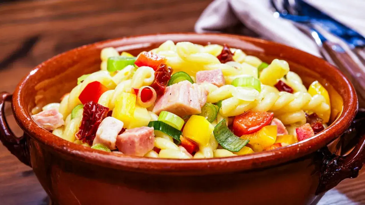 Leckerer nudelsalat ohne mayonnaise mit bunten Paprikawürfeln, Schinken und Frühlingszwiebeln in einer Tonschüssel.