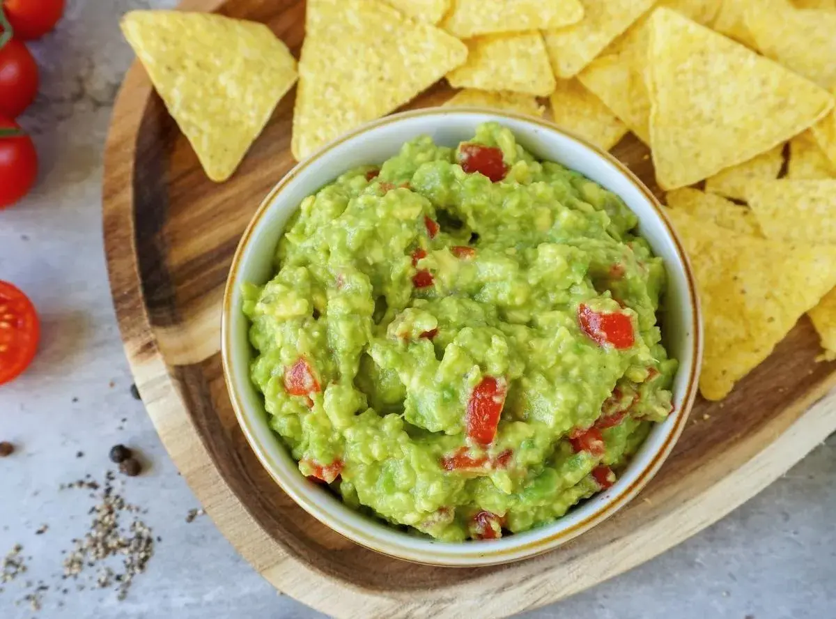 Frisches Guacamole-Rezept mit Tomatenstückchen, serviert mit Tortilla-Chips auf einem Holzbrett.