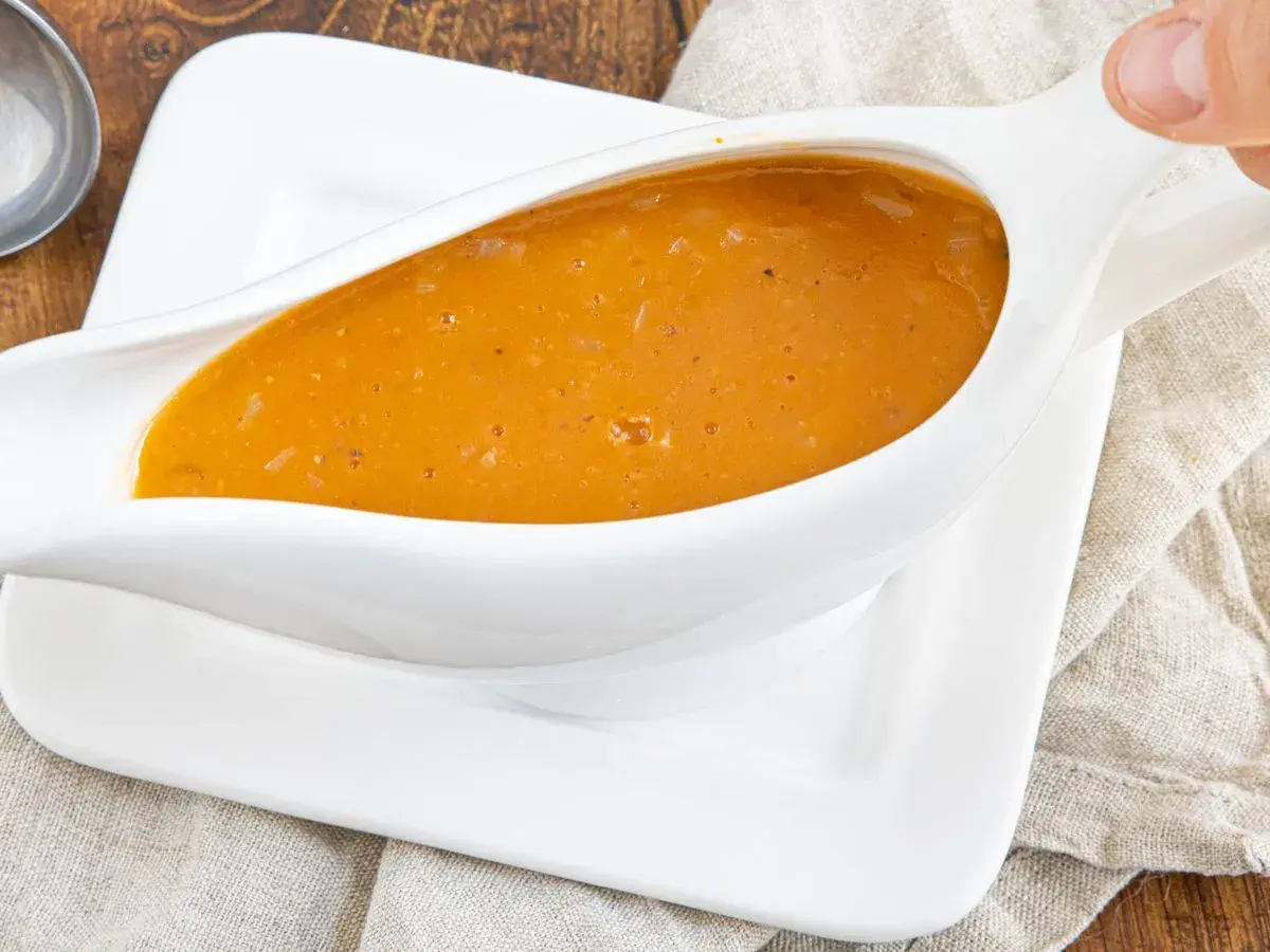 Eine köstliche, goldbraune Sauce mit Fond aus dem Glas, serviert in einer weißen Sauciere auf einem Leinentuch.