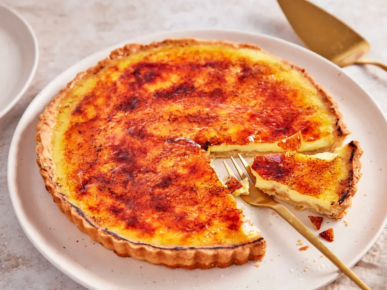 Eine cremige Crème brûlée Tarte mit karamellisierter Zuckerkruste, ein Stück wurde mit einer goldenen Gabel entnommen.