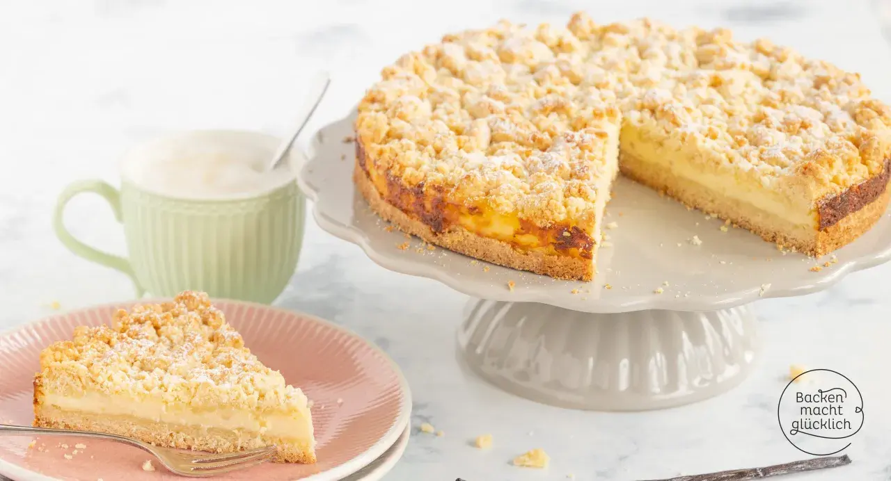 Ein saftiger Streuselkuchen mit Puddingfüllung, bestäubt mit Puderzucker, wartet auf Genießer. Daneben eine Tasse Cappuccino.