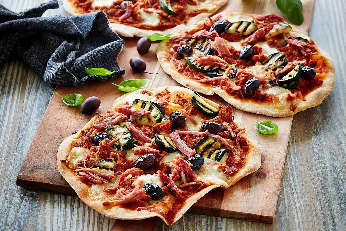 Drei kleine Pizzen mit pulled pork, gegrillter Zucchini und Oliven auf einem Holzbrett.