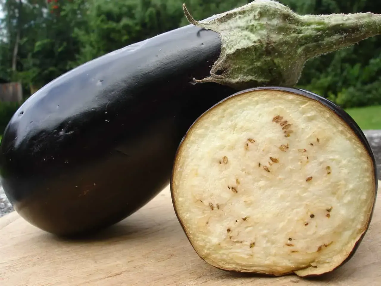 Eine glänzende, dunkle Aubergine liegt neben einem aufgeschnittenen Exemplar. Im Inneren der Aubergine sind viele kleine, braune Kerne zu sehen, die wie schwarze Punkte wirken.