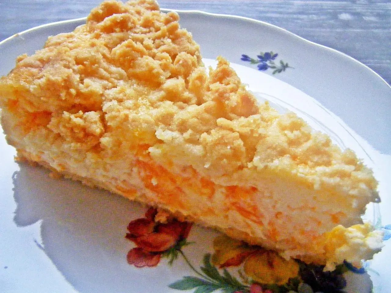 Ein Stück Mandarine Schmand Kuchen mit Streuseln auf einem Teller mit Blumenmuster.
