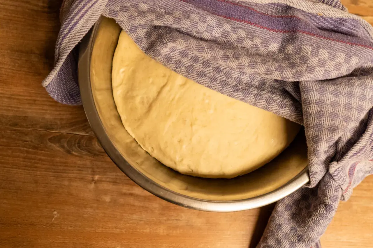 Frischer, aufgegangener Teig für pizzateig selber machen ruht in einer Schüssel unter einem karierten Tuch.