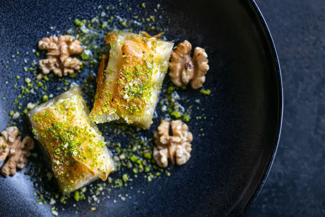 Zwei goldbraune Baklava-Stücke, bestreut mit gehackten Pistazien und Nüssen, auf einem dunklen Teller. Ein köstliches Baklava Rezept.