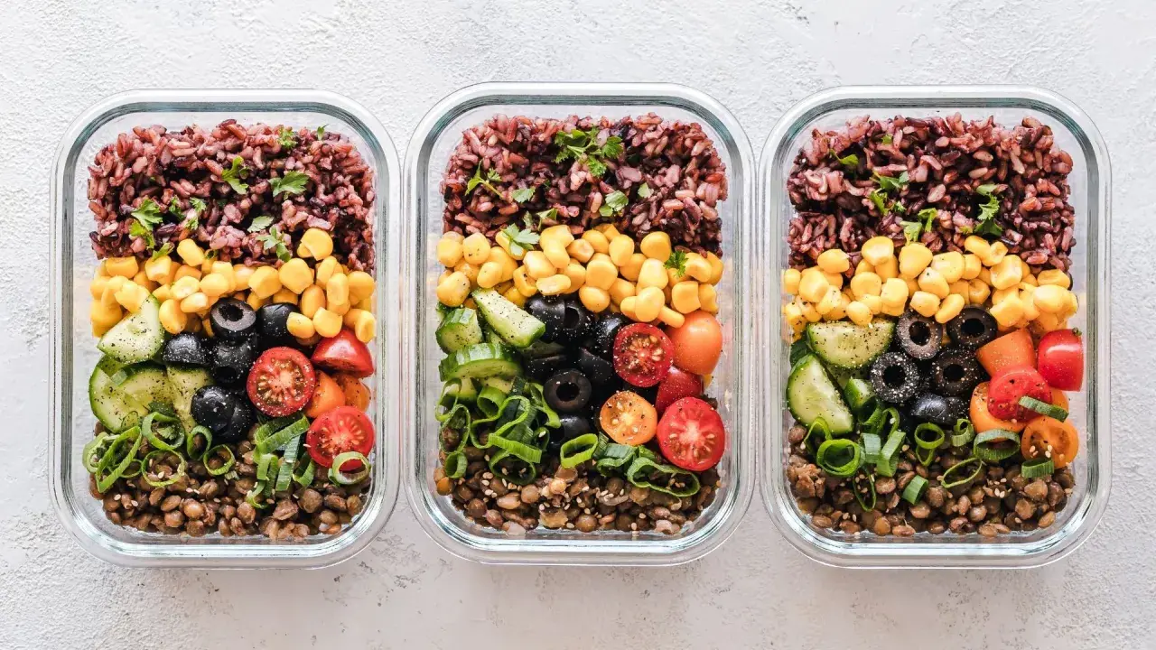 Drei Glasbehälter mit Linsen, Gurken, Tomaten, Oliven, Mais und braunem Reis – tolle meal prep ideen für gesunde Mahlzeiten.