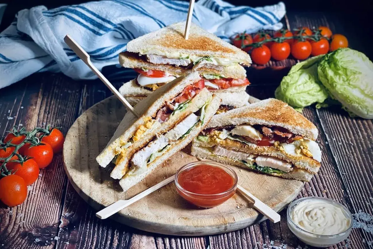 Drei saftige Club Sandwiches mit Hähnchen, Speck, Ei und Tomaten, serviert mit Ketchup und Mayo.
