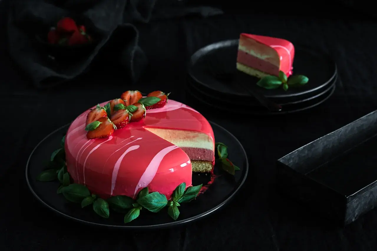Eine Mousse Torte mit glänzendem Spiegelglasur, dekoriert mit Erdbeeren und Basilikum. Ein Stück ist auf einem Teller angerichtet.