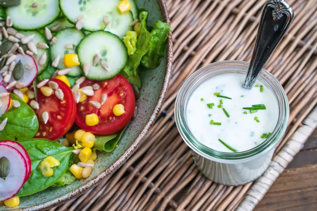 Frischer Salat mit Tomaten, Gurken und Mais neben einem Glas mit selbstgemachtem Joghurt-Dressing, garniert mit Schnittlauch.