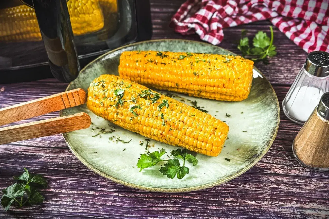Zwei goldgelbe Maiskolben, frisch aus dem Airfryer, mit Kräutern bestreut, liegen auf einem Teller. Daneben Salz- und Pfefferstreuer.