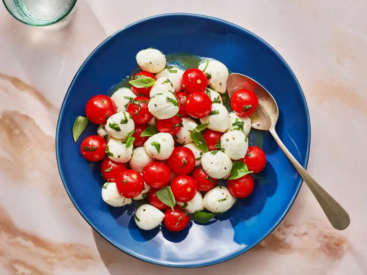 Ein bunter italienischer Salat mit Kirschtomaten, Mini-Mozzarella-Kugeln und frischem Basilikum auf einem blauen Teller.