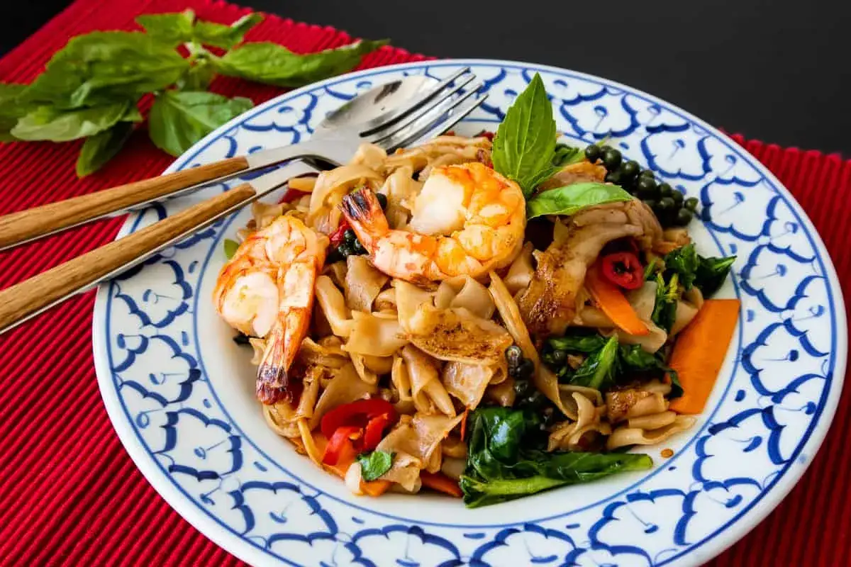 Pad Kee Mao mit Garnelen, breiten Reisnudeln, Gemüse und frischem Basilikum auf einem Teller mit blau-weißem Muster.