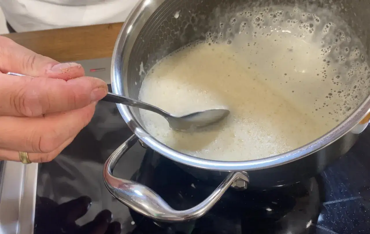 Eine Hand rührt mit einem Löffel in einer Pfanne mit schäumender Flüssigkeit, die nach beurre blanc aussieht.