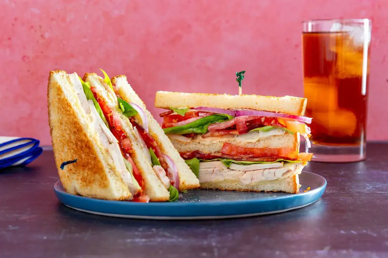Ein saftiger Club Sandwich mit Hähnchen, Speck, Tomaten und Salat, daneben ein Glas Eistee.