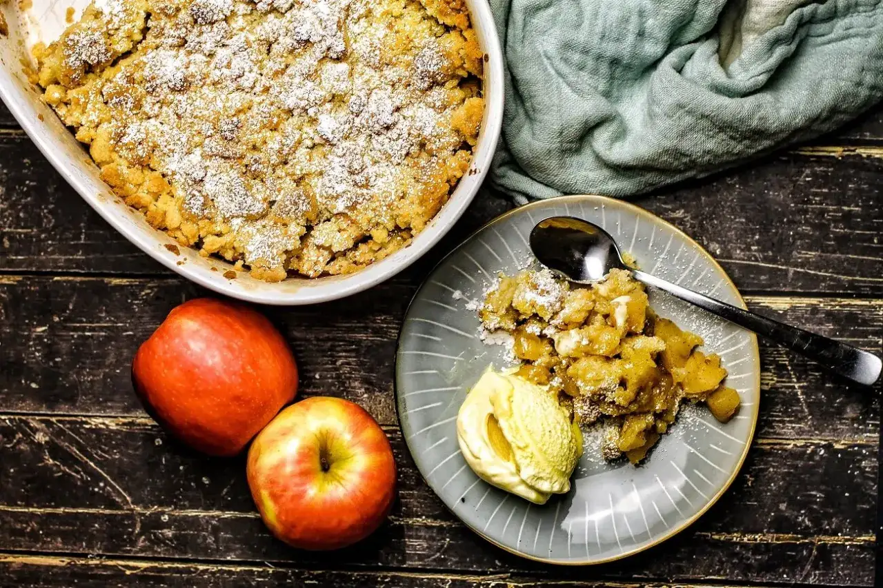Ein goldbrauner Apfelcrumble mit Puderzucker bestreut, daneben eine Kugel Vanilleeis auf einem Teller. Zwei Äpfel liegen daneben.