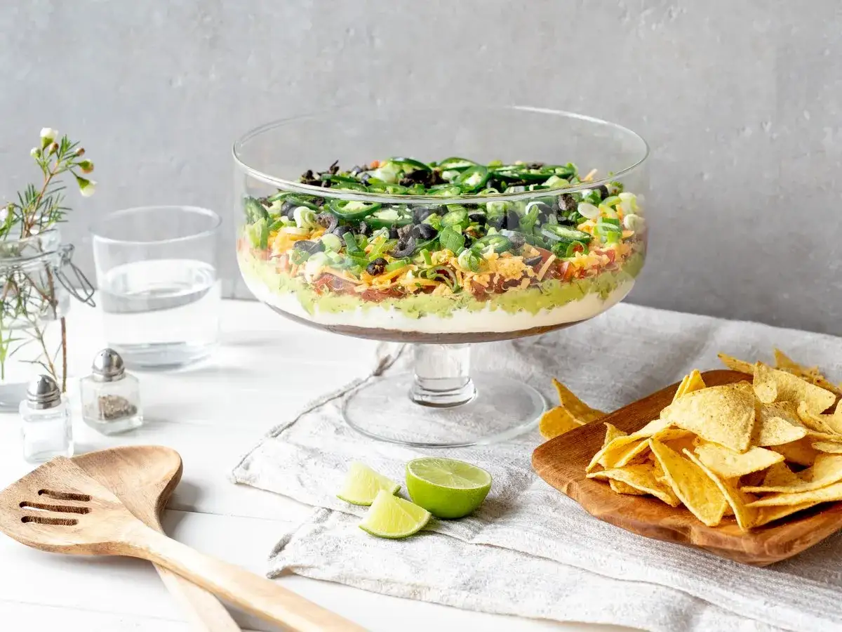 Schicht für Schicht ein Genuss: Ein bunter 7-Layer-Dip in einer Glasvitrine, bereit zum Dippen mit Tortilla-Chips.