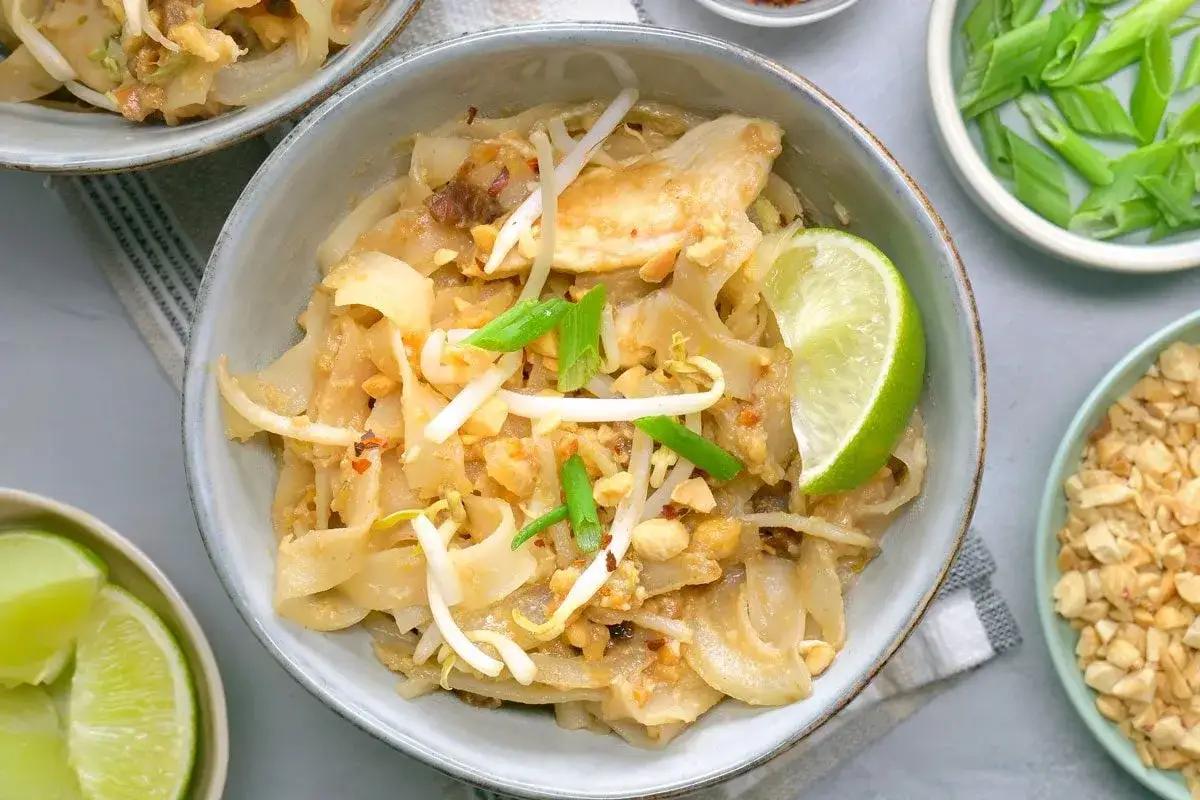 Sch&uuml;ssel mit Pad Thai Gai, garniert mit Limettenspalte, Sojasprossen und Fr&uuml;hlingszwiebeln. Daneben gehackte Erdn&uuml;sse.