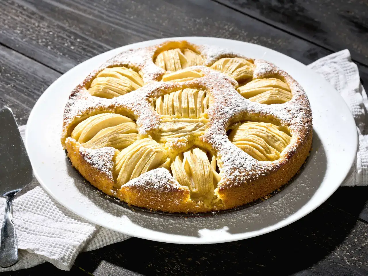 Ein goldbrauner Apfelkuchen, bestäubt mit Puderzucker, wartet auf sein apfelkuchen rezept. Daneben liegt ein Tortenheber.