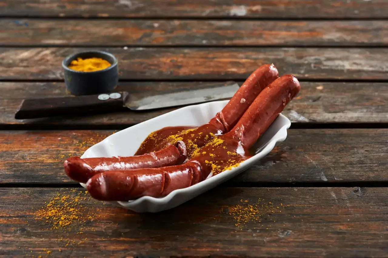 Drei Bratw&uuml;rste in einer wei&szlig;en Schale mit einer dunklen Sauce und Currypulver. Ein kleiner Napf mit Currypulver und ein Messer liegen daneben. Das ist das rezept currywurst sauce original.