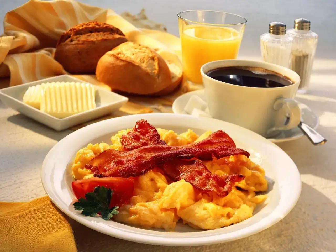 Ein herzhaftes full english breakfast mit Rührei, Speck, Tomate, Brötchen, Butter, Orangensaft und Kaffee.