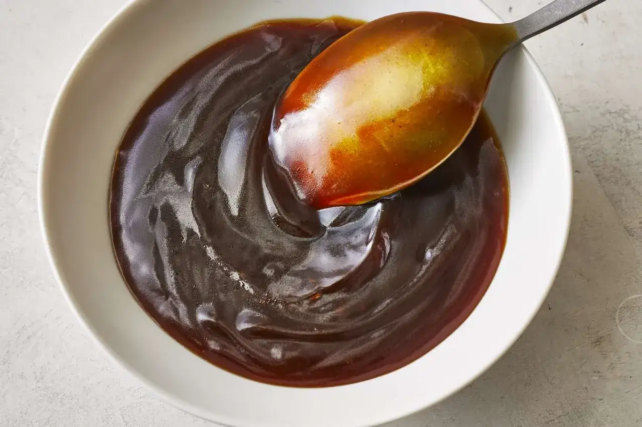 Eine L&ouml;ffelspitze hebt gl&auml;nzende, dunkle Teriyaki-Sauce aus einer wei&szlig;en Sch&uuml;ssel. Was ist Teriyaki? Eine s&uuml;&szlig;-salzige Glasur f&uuml;r Fleisch und Fisch.
