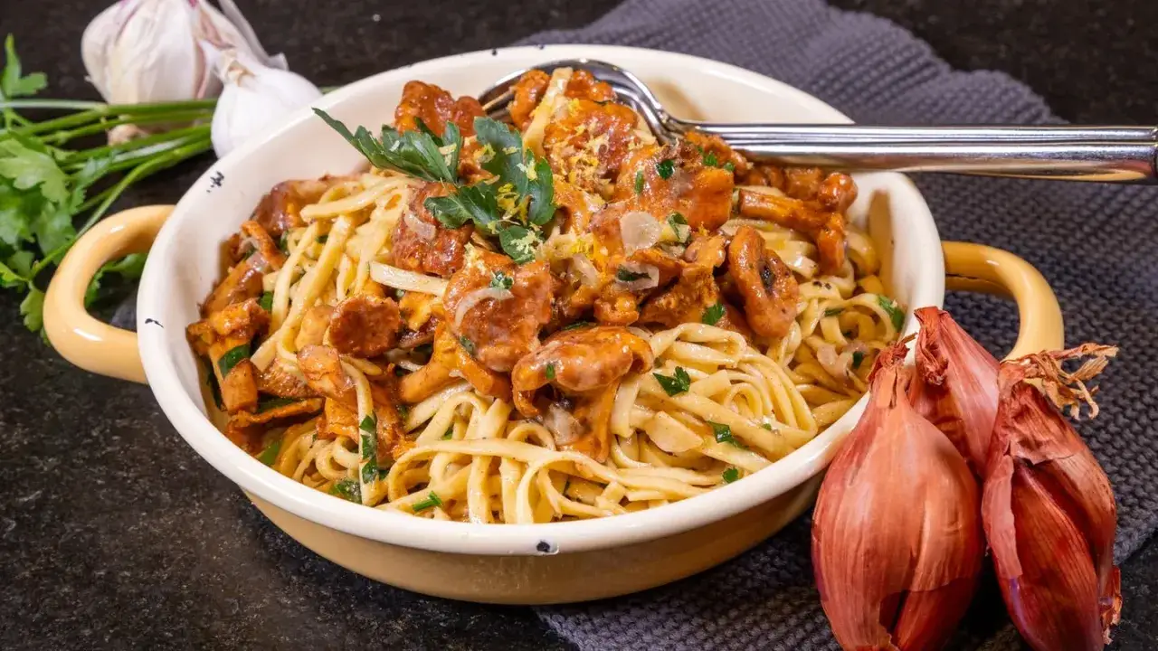 Tagliatelle mit Pfifferlingen, garniert mit Petersilie und Zitronenschale. Daneben liegen Schalotten und Knoblauch.
