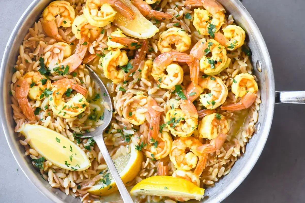 Orzo mit Garnelen in einer Pfanne, garniert mit Zitronenspalten und Petersilie. Ein Löffel hebt eine Portion hervor.
