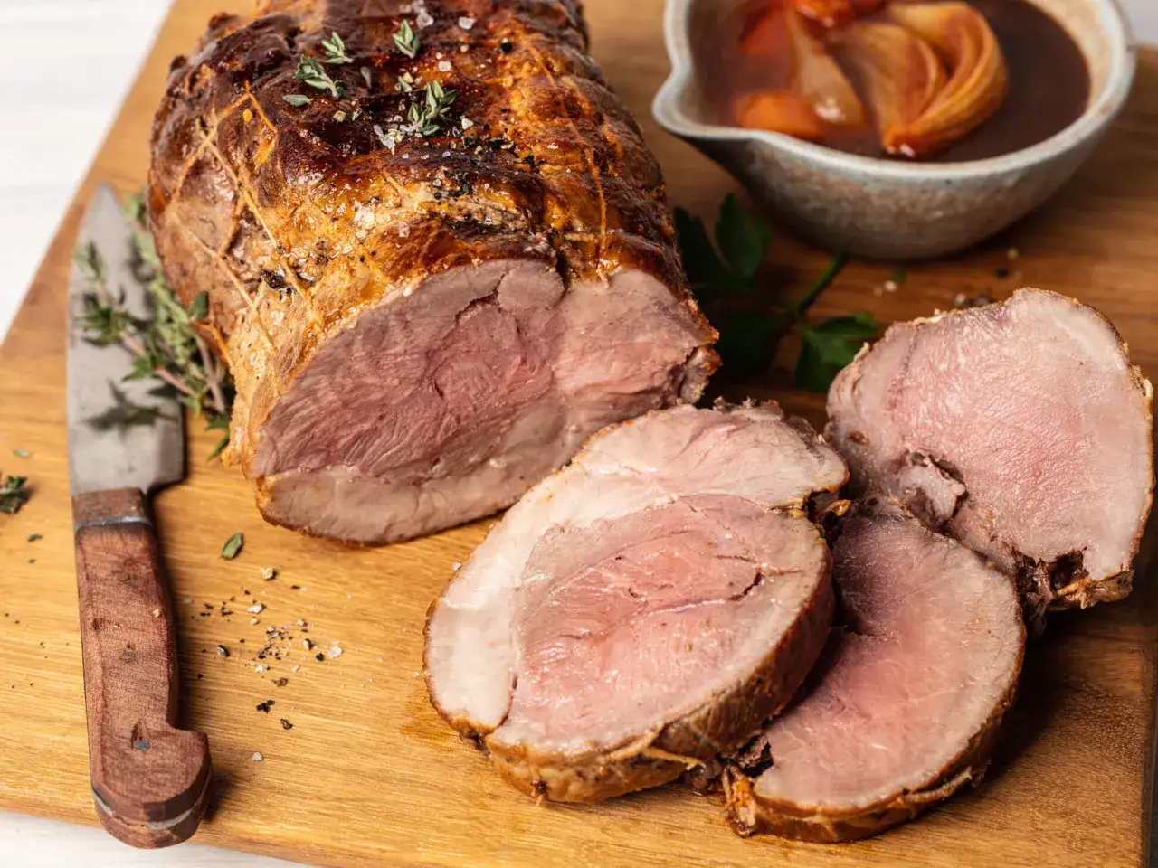 Saftiger Rollbraten im Backofen auf Holzbrett, mit Kr&auml;utern und einer Sch&uuml;ssel Sauce.
